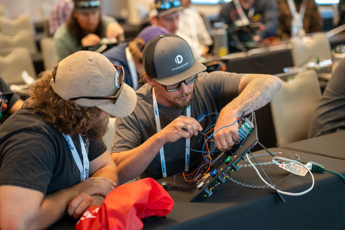 Two men working on electrical wiring at a workshop table.