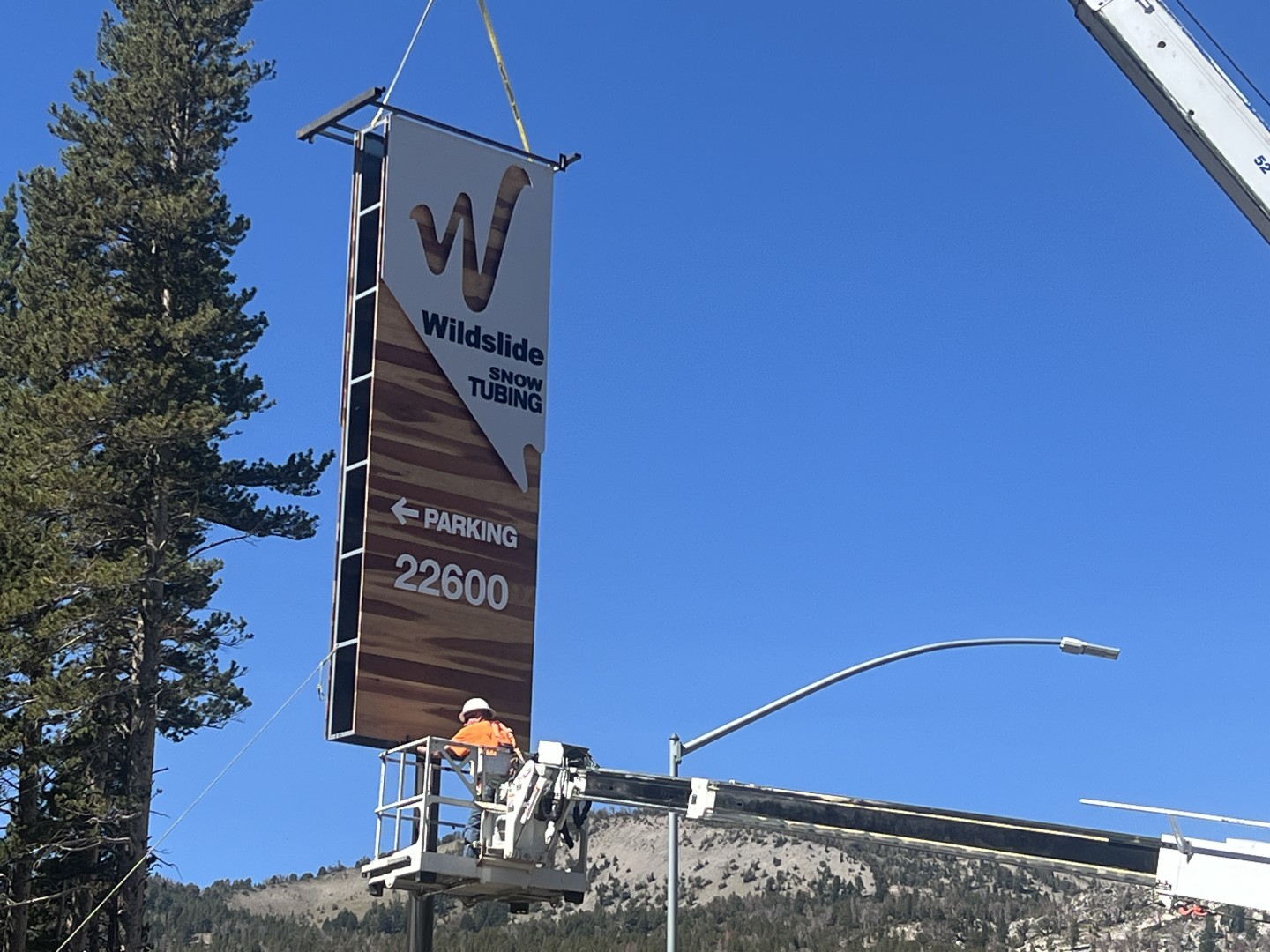 Sign on crane near tree and mountains, under a clear blue sky.
